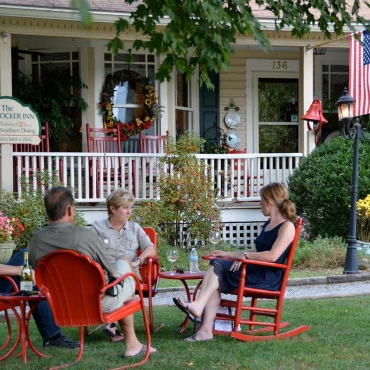People socializing on a lawn in front of a yellow house with a white porch. They sit in red chairs around a small table.