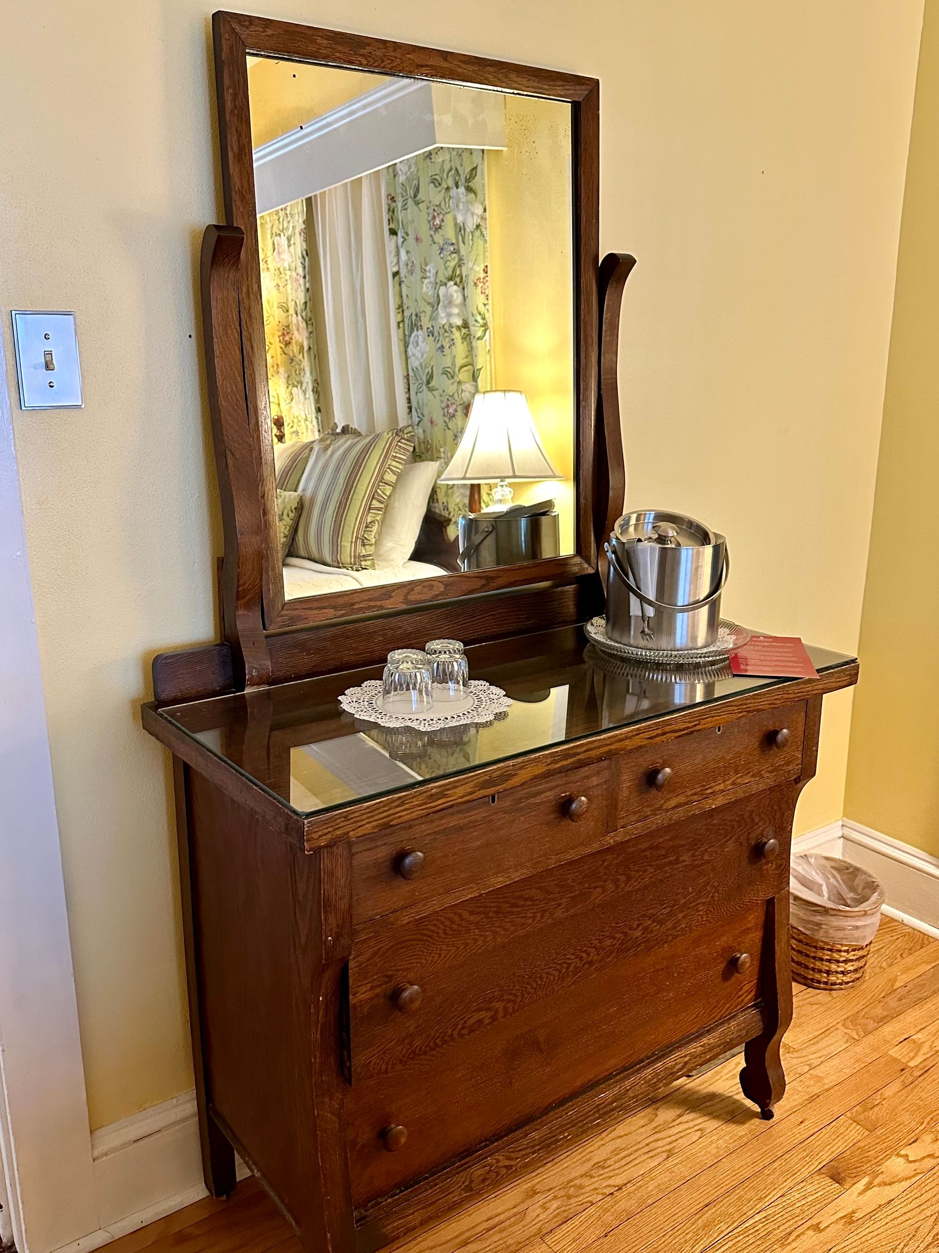 Wooden dresser with mirror reflecting a bed, lamp, and other room details.