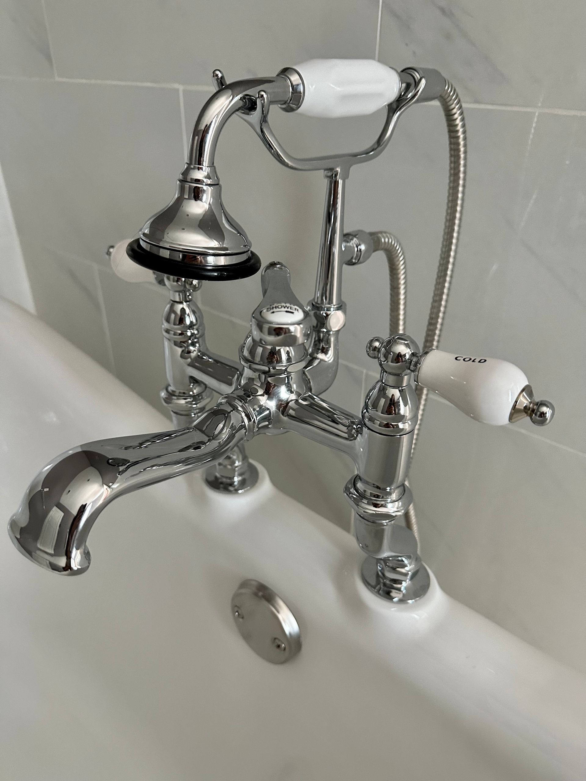 Chrome bathtub faucet with a hand shower and white ceramic handles. Set against gray tiled wall.
