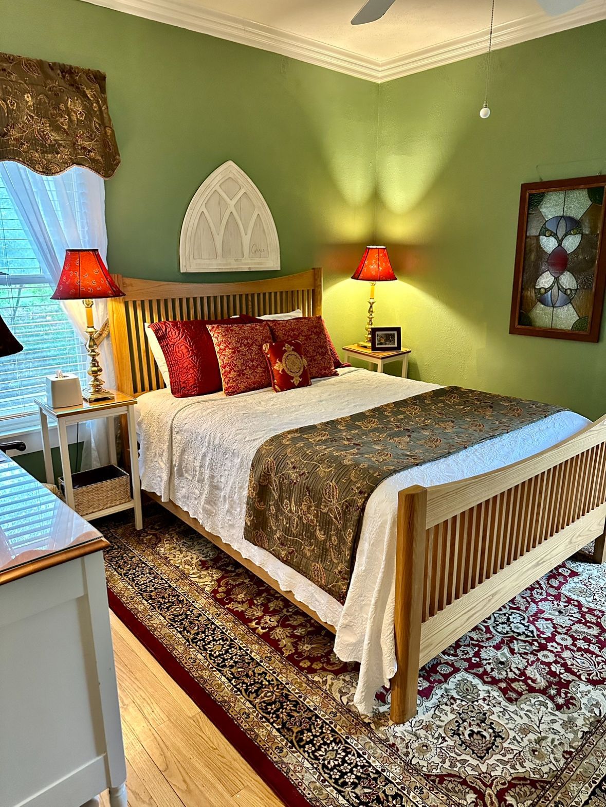 A bedroom with a light-colored wooden bed, red lamps, and green walls. The bed is made with red pillows and a patterned blanket.