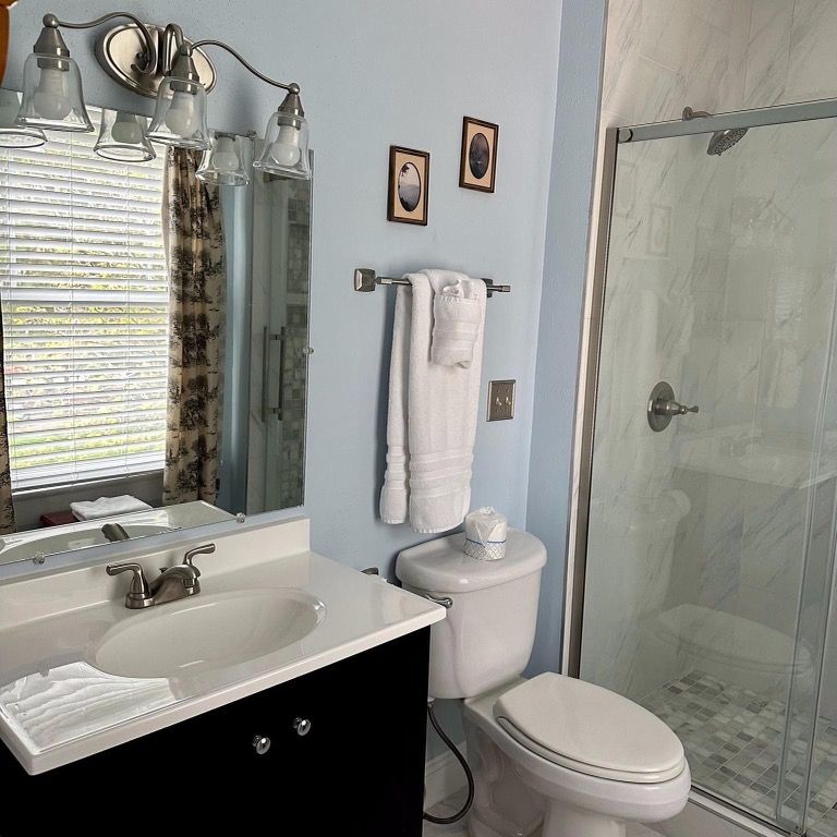 A bathroom with a shower, toilet, vanity, and window. Light blue walls, a black vanity, and white fixtures.