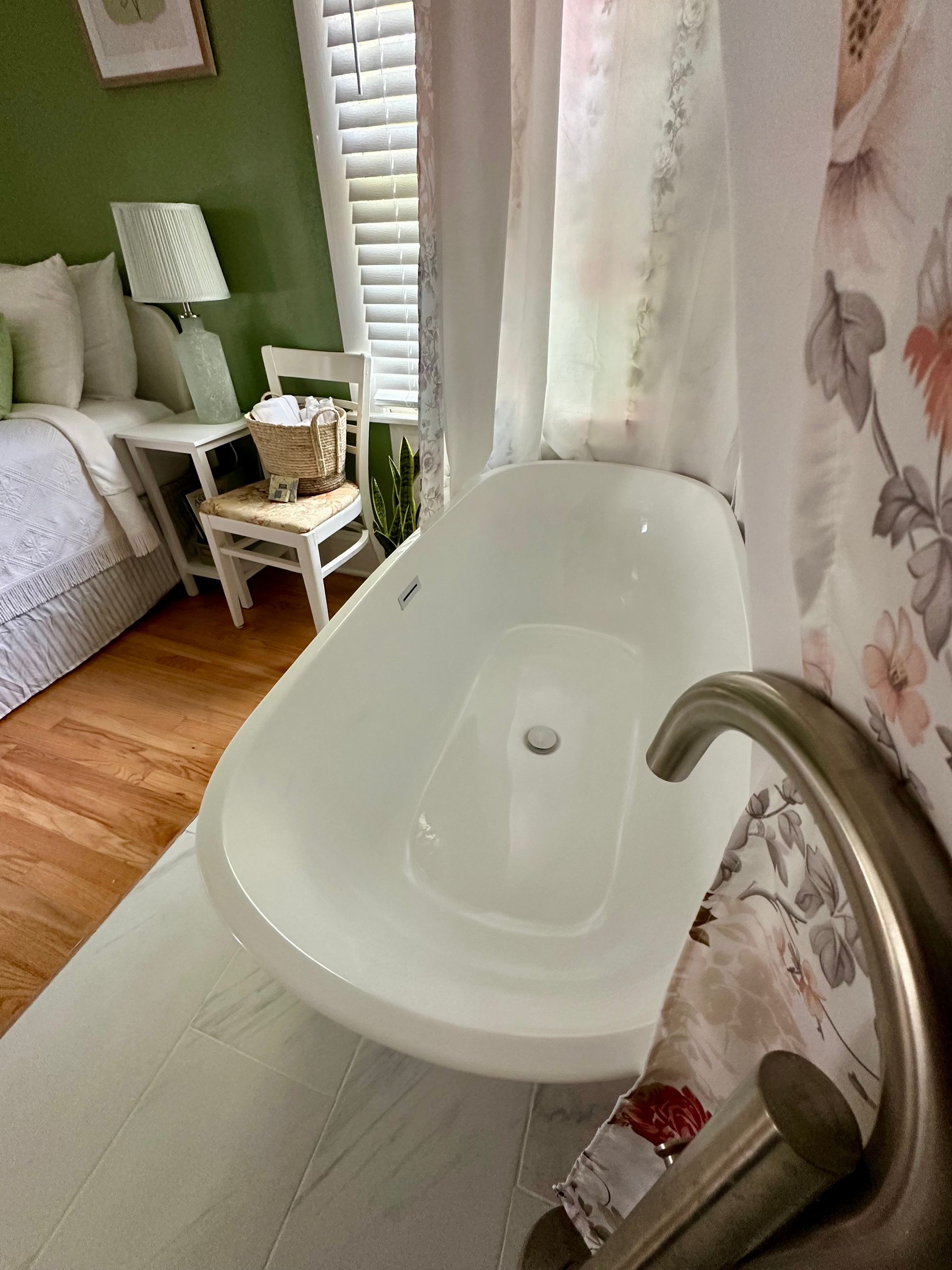 Clawfoot bathtub in a room with a bed, chair, and window. The room is green, with white curtains and wooden floors.
