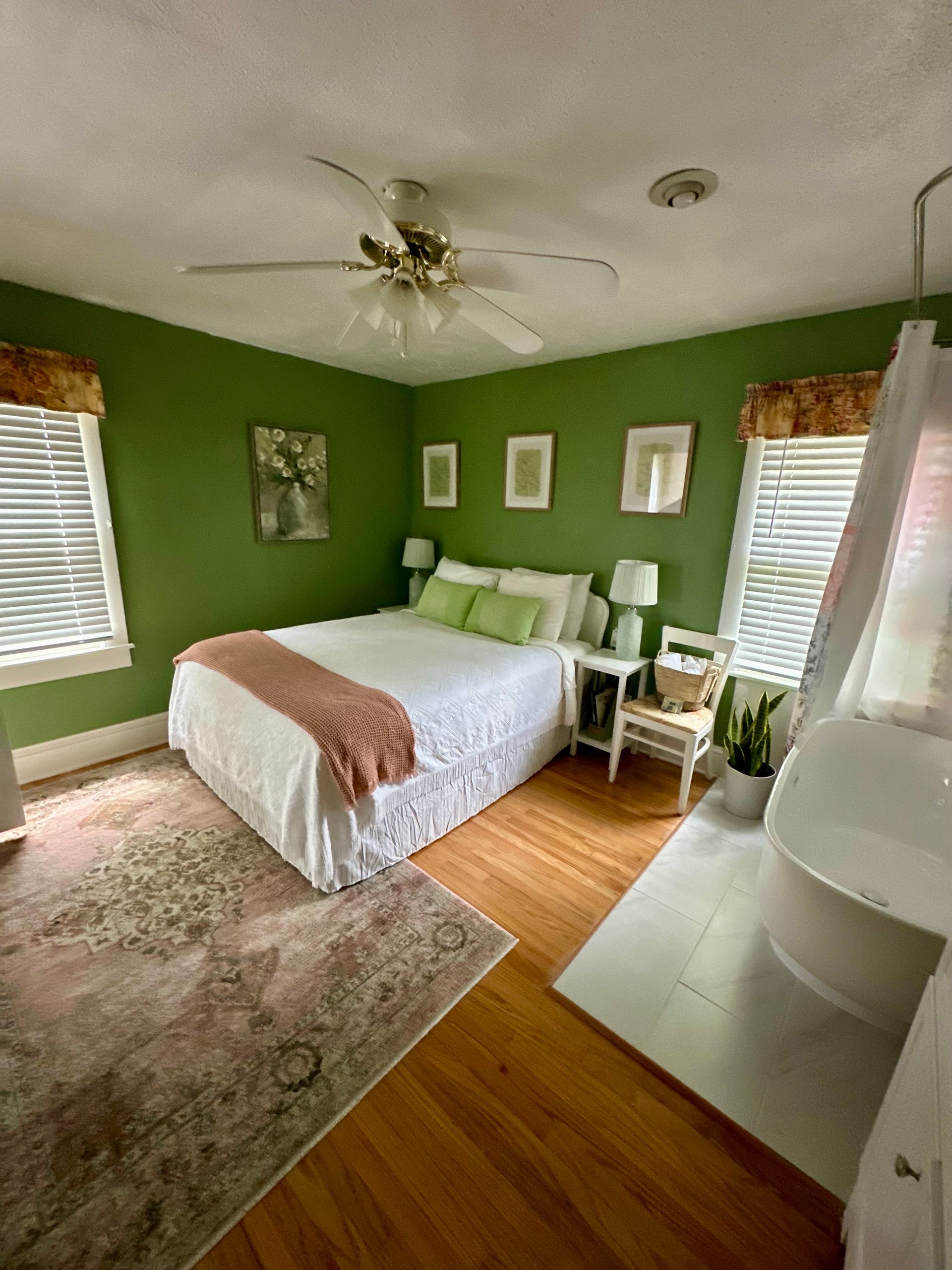 Green-walled bedroom with a white bed, framed artwork, and a large rug. A fan hangs from the ceiling, and light shines through the blinds.