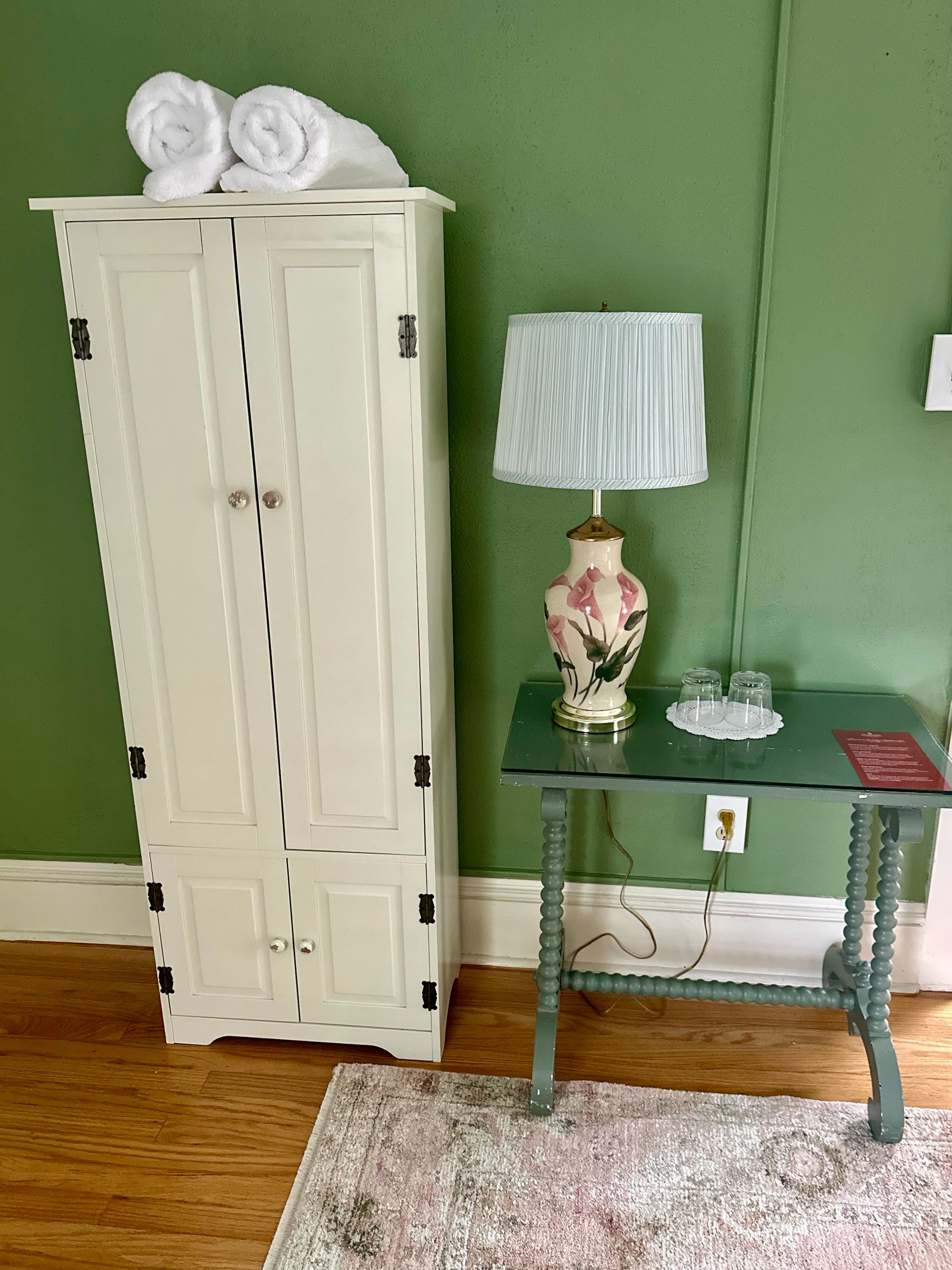 A cream-colored cabinet with rolled towels on top next to a small table with a lamp and glasses against a green wall.