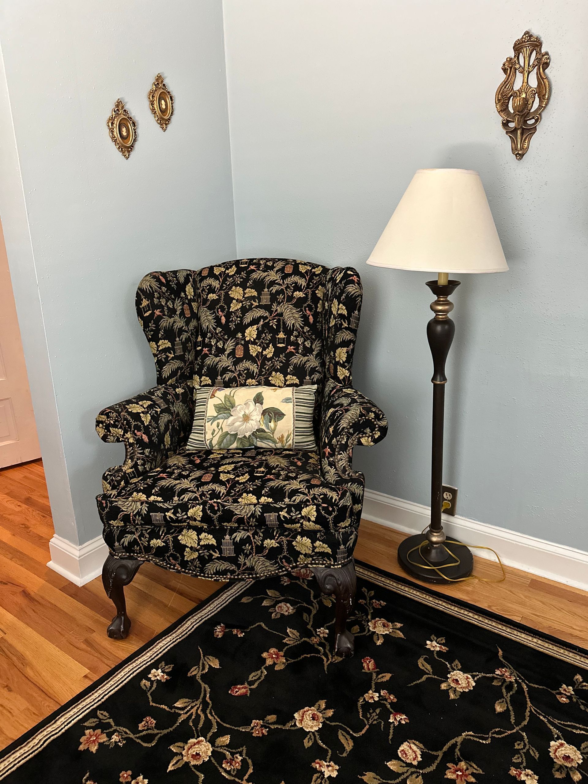 A floral armchair and lamp in a corner, set on a patterned rug against a light blue wall with sconces.