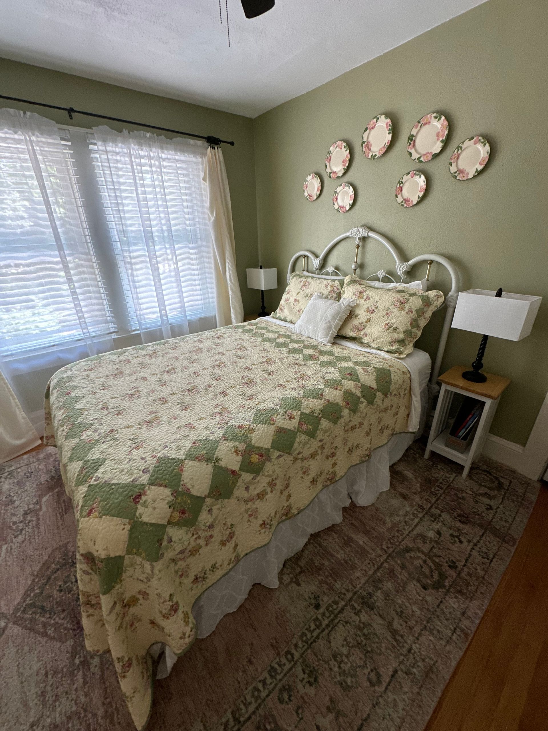 Bedroom with a white iron bed, floral quilt, armchair with a red throw, and pale green walls. Decorative plates hang on the wall.