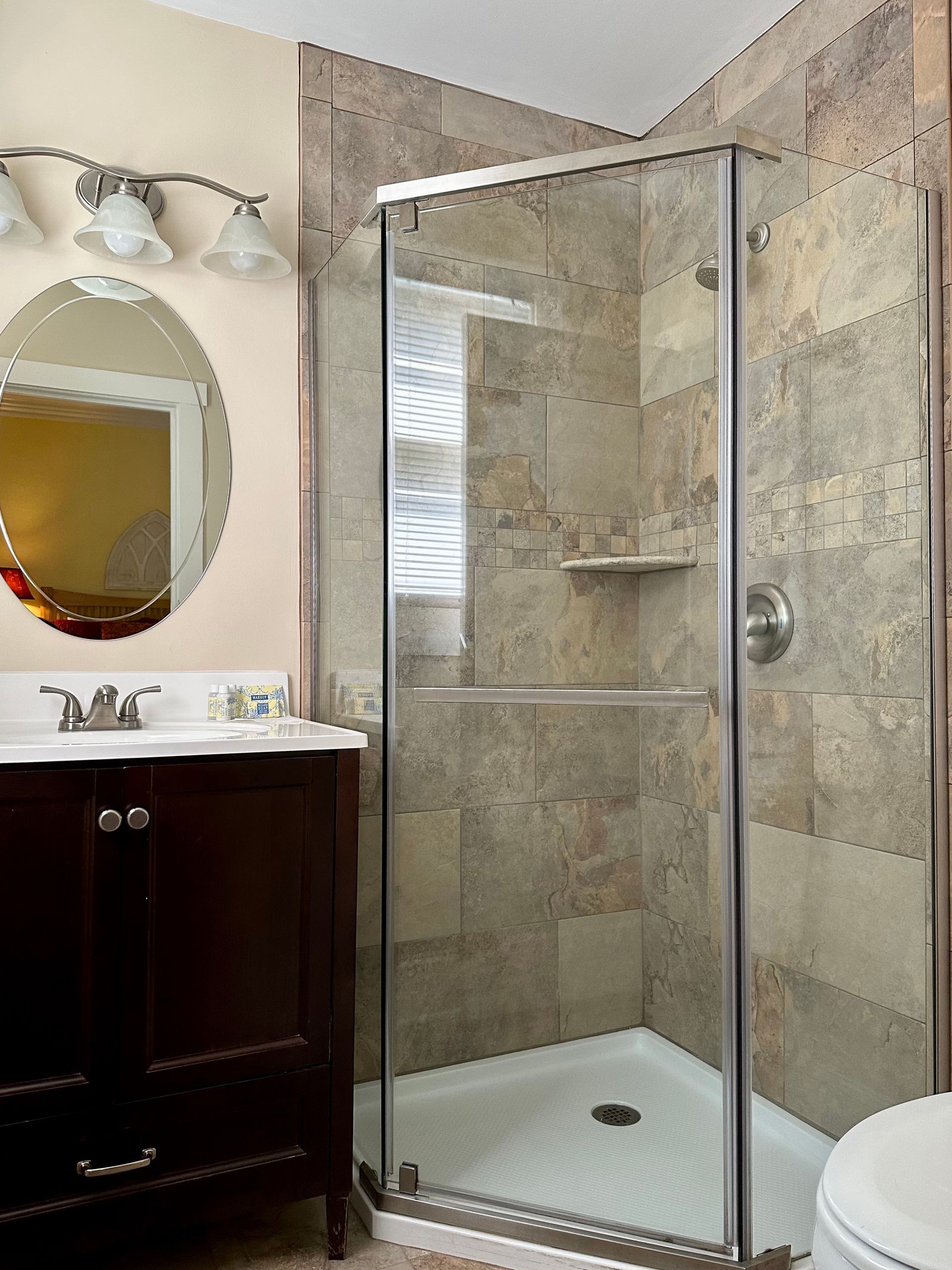 Bathroom with a glass shower, dark wood vanity, oval mirror, and tan tile walls.