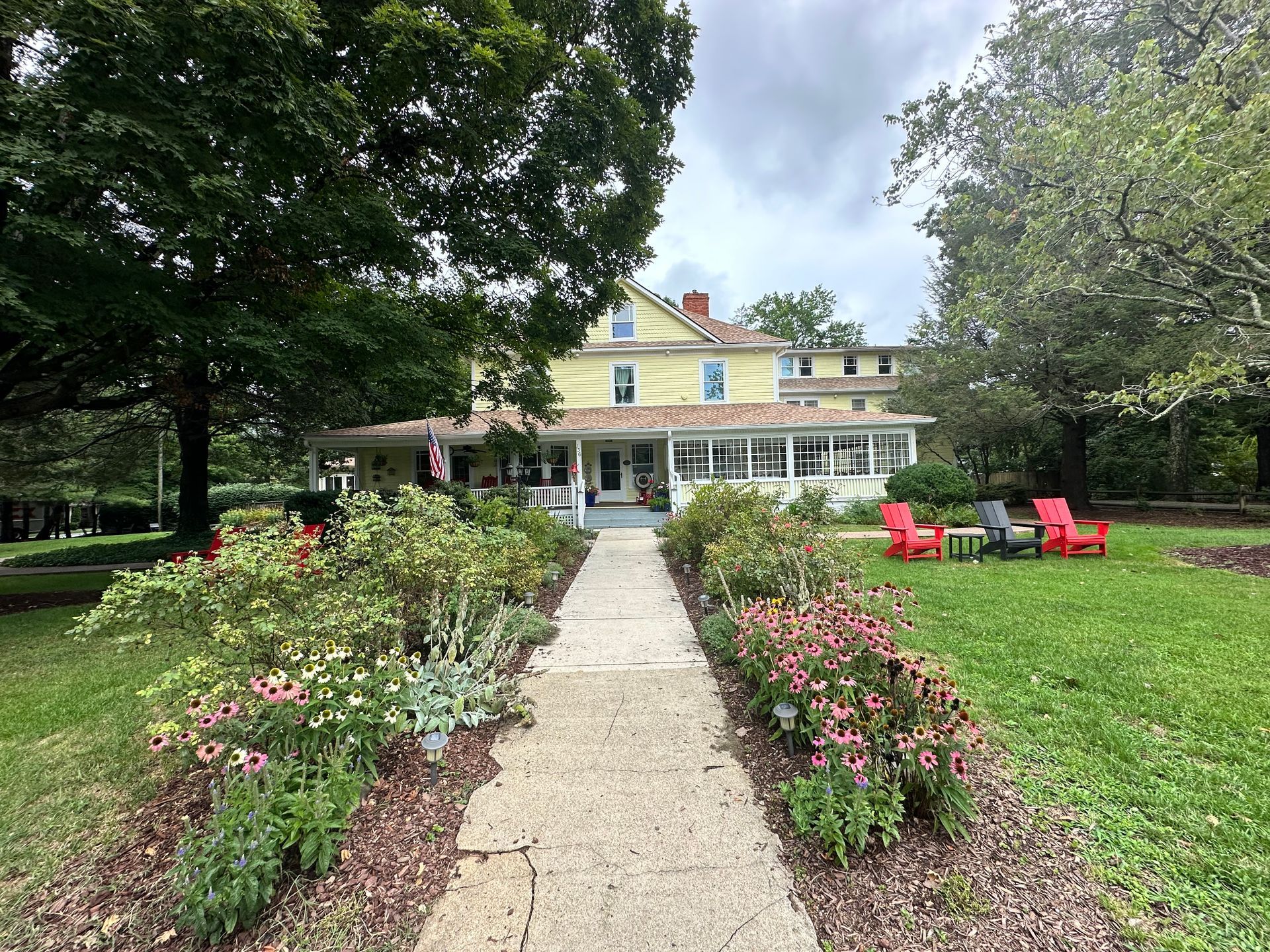 Two-story house with a wrap-around porch and green lawn. Trees frame the house with a blue and white striped awning over the porch.