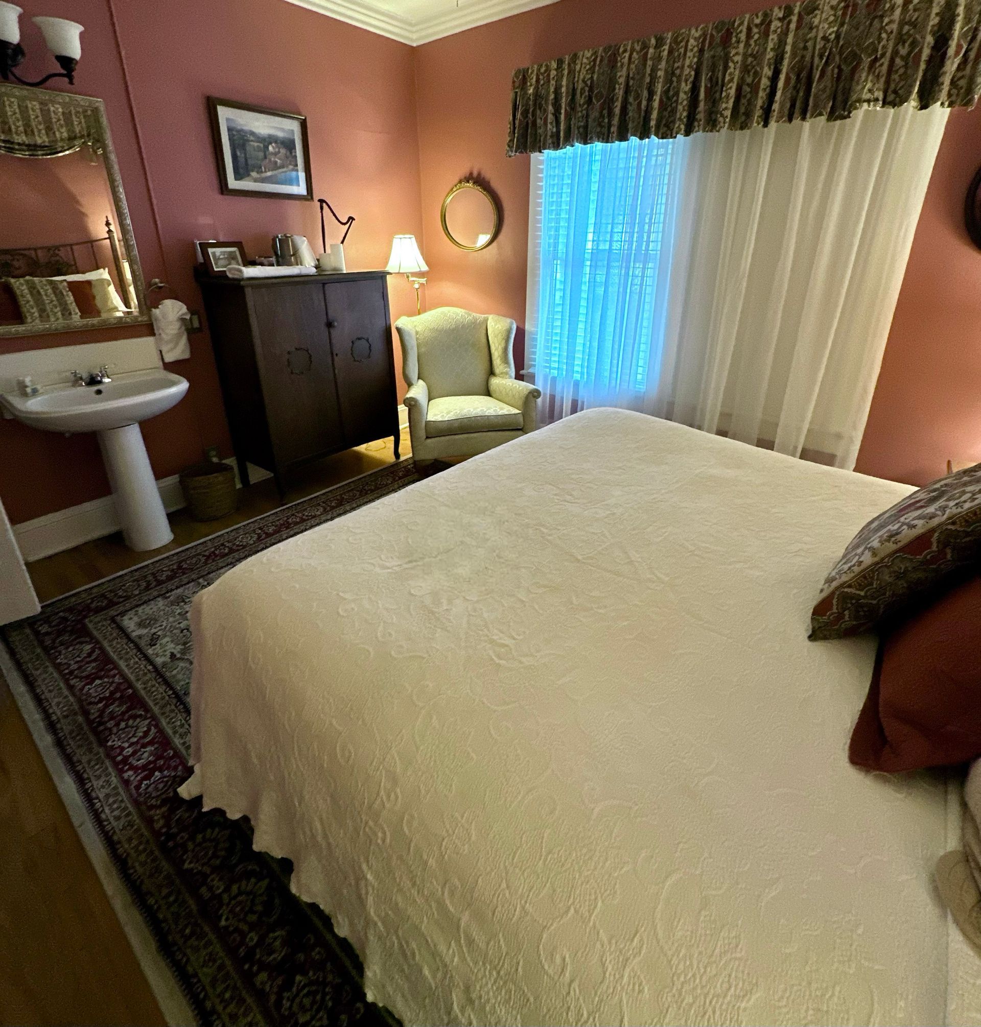 A cozy bedroom with mauve walls, hardwood floors, and antique furnishings, including a vanity, armoire, and armchair.