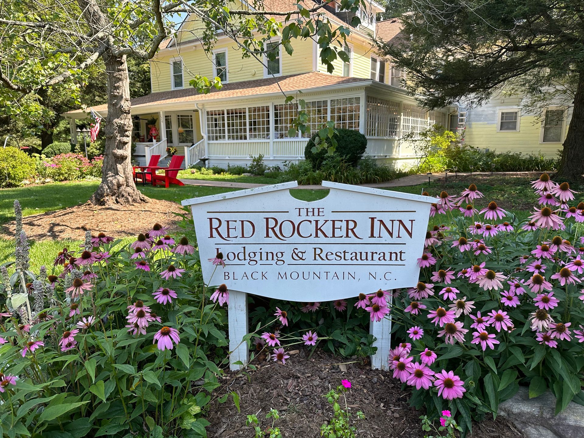 A house front with a white porch, red chairs, and a vintage fire alarm bell. Purple rhododendrons bloom in the foreground.