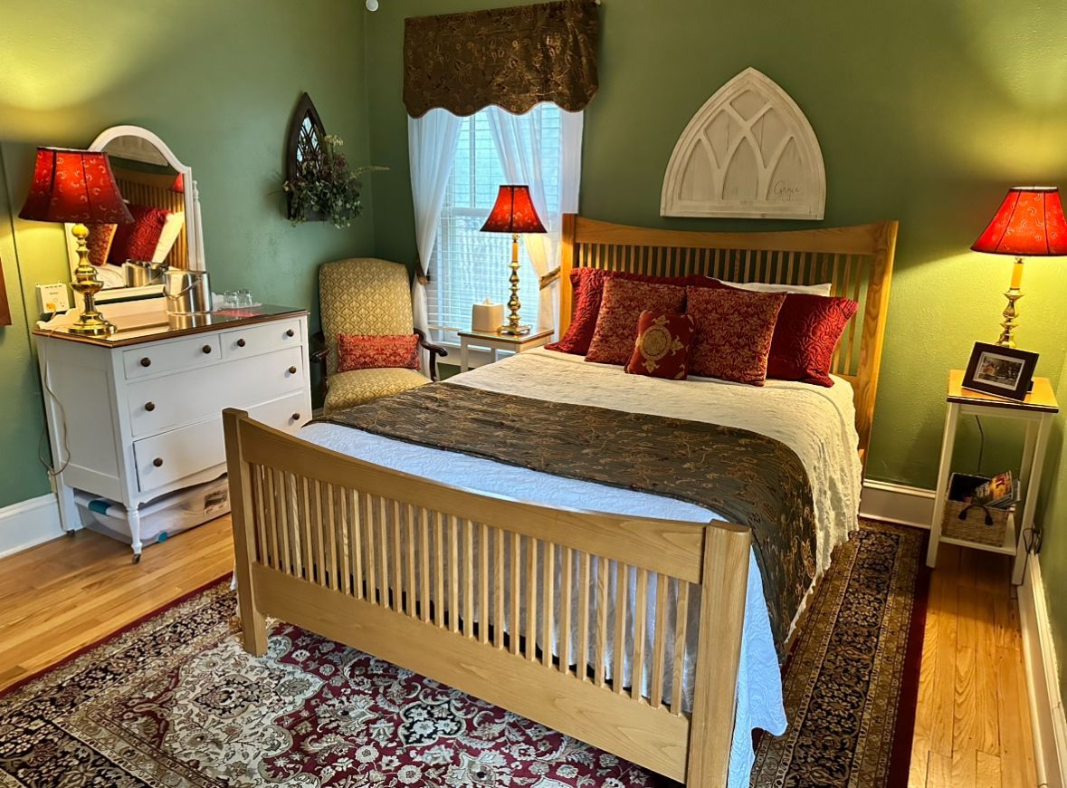 A bedroom with a light-wood bed, white dresser, and green walls. The bed has red pillows and a brown runner. Lamps with red shades flank the bed.