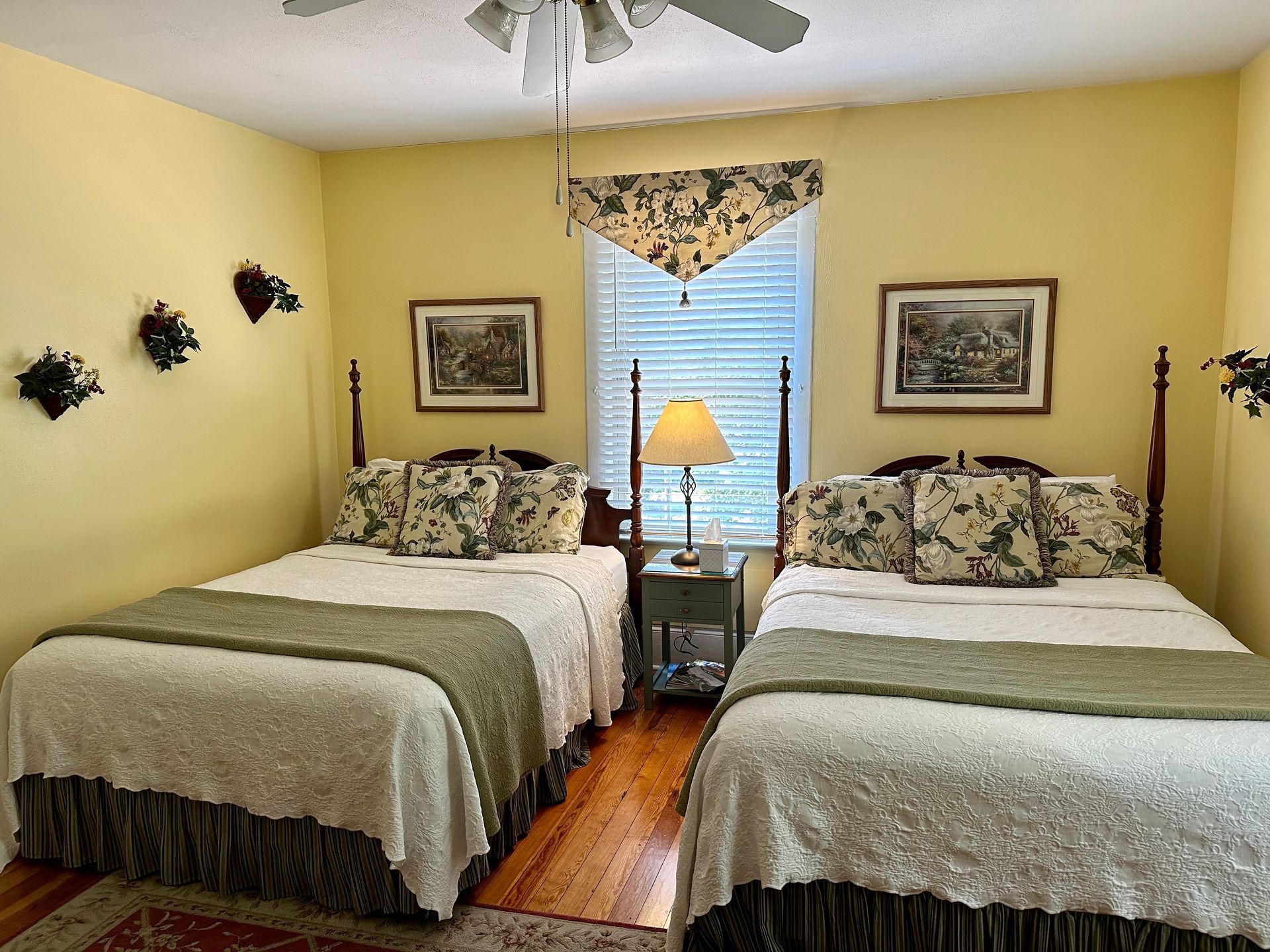Two beds in a yellow room with floral bedding, art, and a window. A lamp sits on a nightstand between the beds.