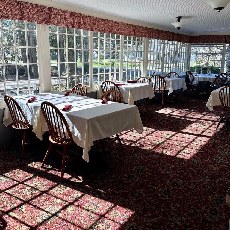 A bright, sunlit dining room with tables set for meals. Windows offer a view of the outdoors, and sunlight casts shadows on the red patterned carpet.