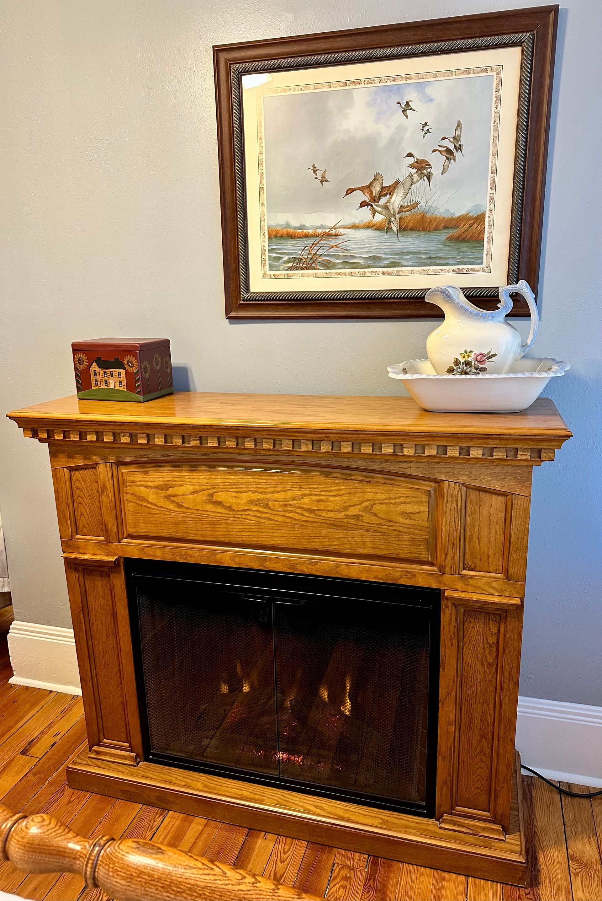 Wooden fireplace mantel with a framed print above, a washbowl, and a small box on top. The room has light blue walls.