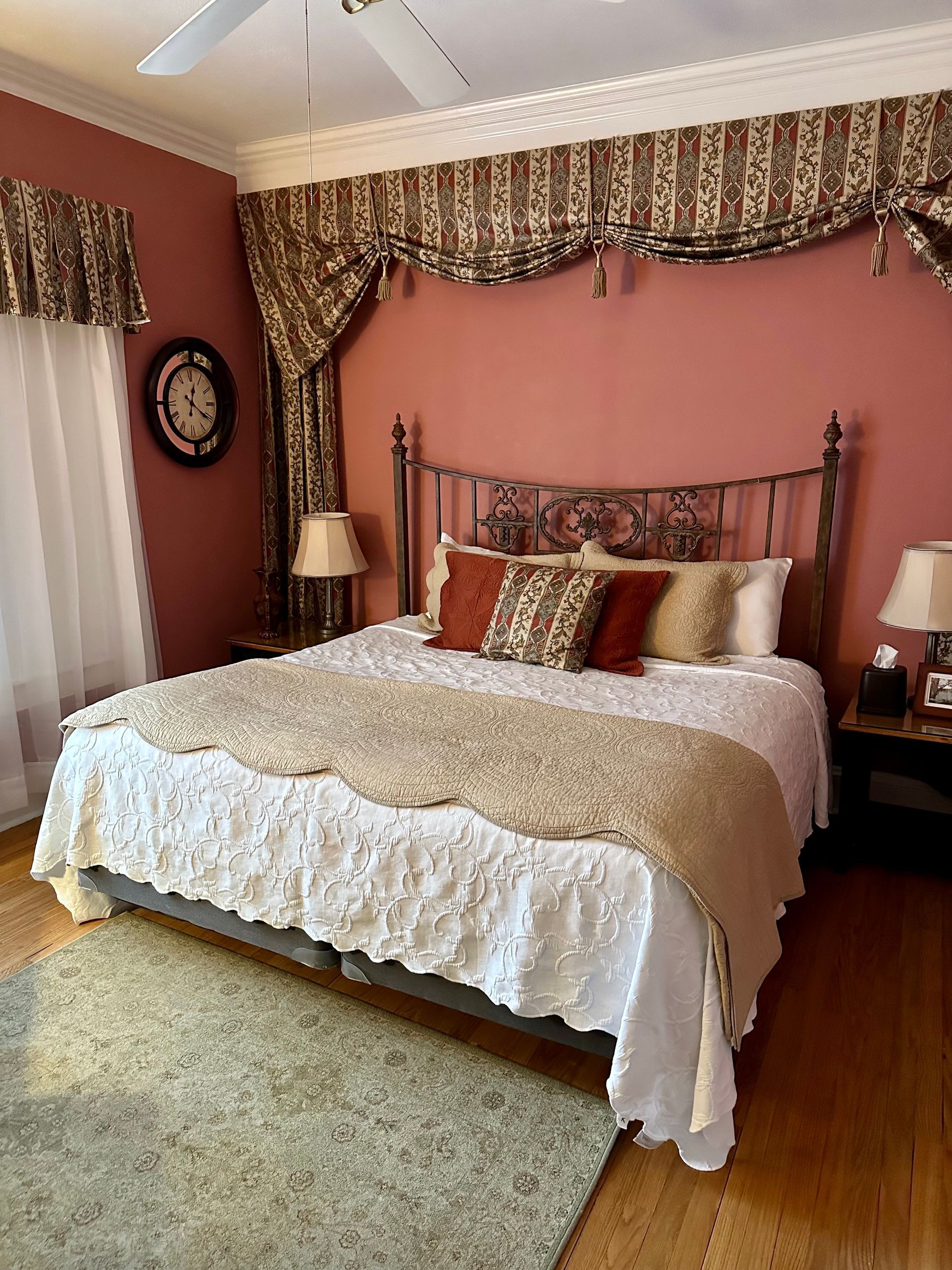 Bedroom with a wrought-iron bed, pink walls, and patterned curtains. A rug and nightstands with lamps complete the cozy space.
