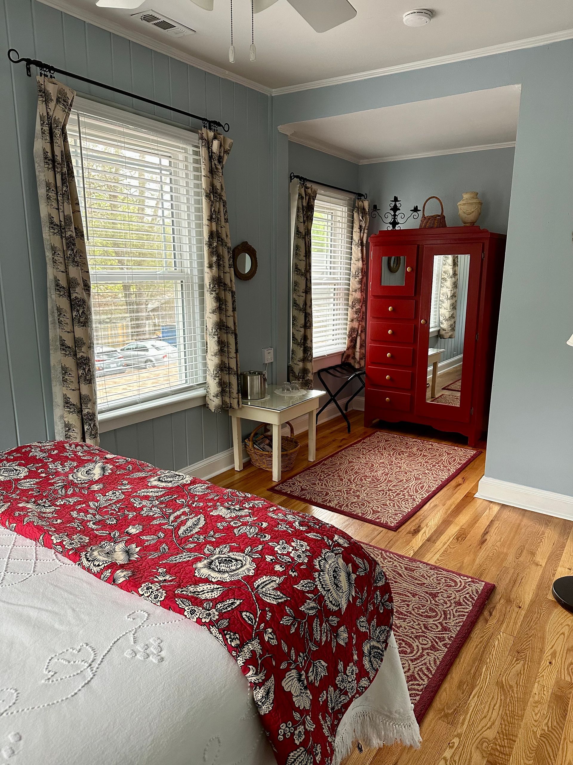 A bedroom with a red bedspread, blue walls, and a red wardrobe. Two windows with floral curtains are visible.
