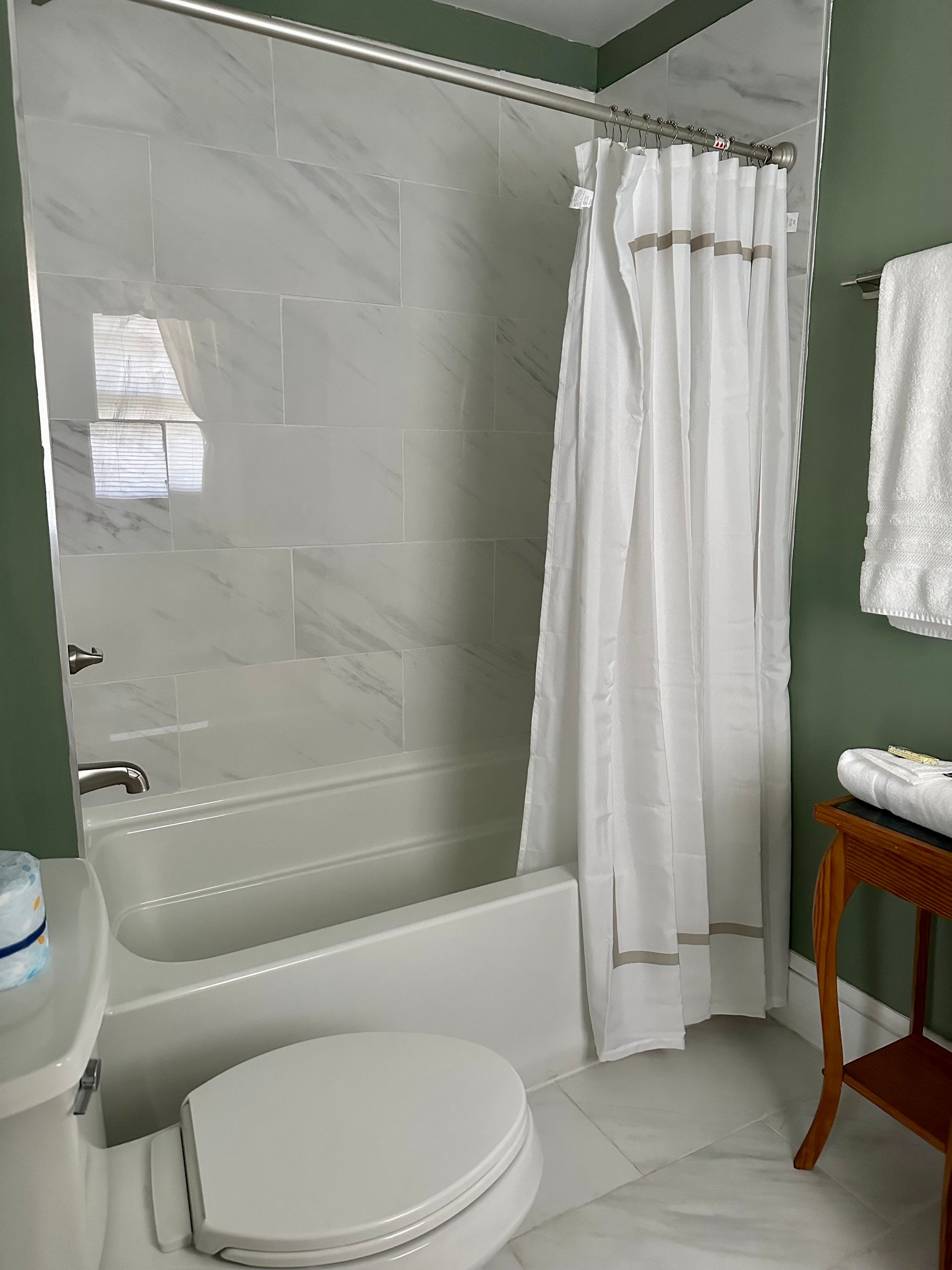 A white bathroom with gray marble-look tiles, a shower curtain, and a toilet. Green walls and a towel rack are on the right.
