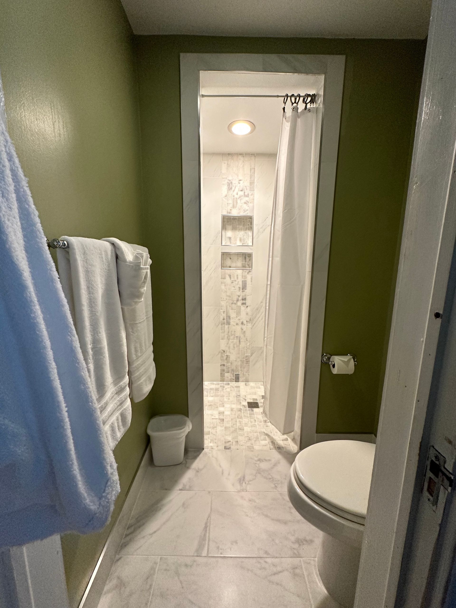 A small bathroom with a white toilet, marble floor, and a shower visible in the back. Green walls, white towels, and a wastebasket are also present.