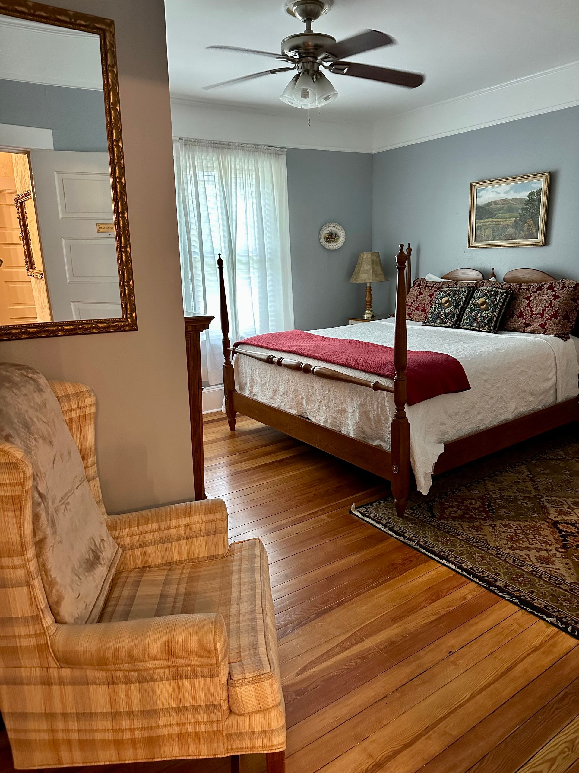 Bedroom with wooden floor, four-poster bed with red blanket, patterned chair, and a large mirror. Pale blue walls.