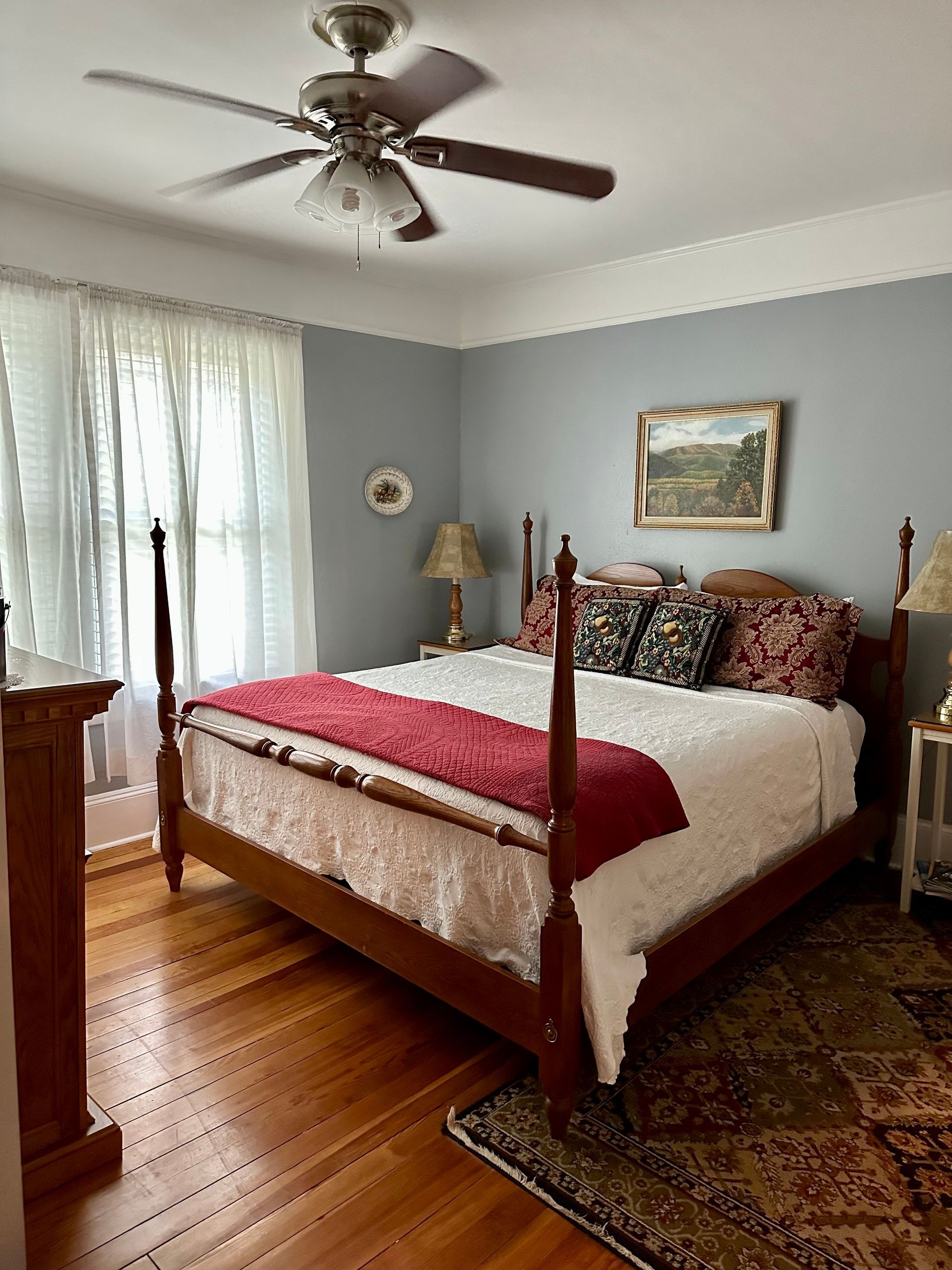 Bedroom with a four-poster bed, hardwood floors, and blue-gray walls. A ceiling fan hangs above the bed, and a window with curtains is on the left.