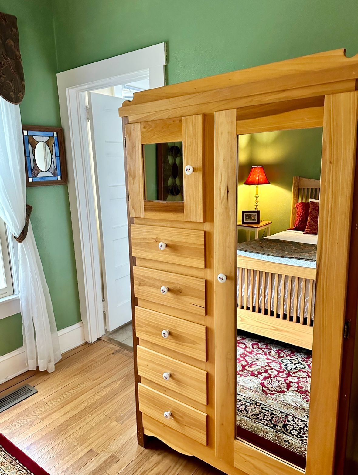 Wooden armoire with a mirror door, reflecting a bed in a bedroom with green walls and a red rug.