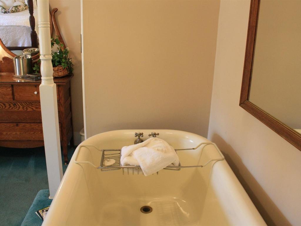 A vintage bathtub with a towel resting on a metal rack, near a mirror and wooden furniture in a light-colored room.