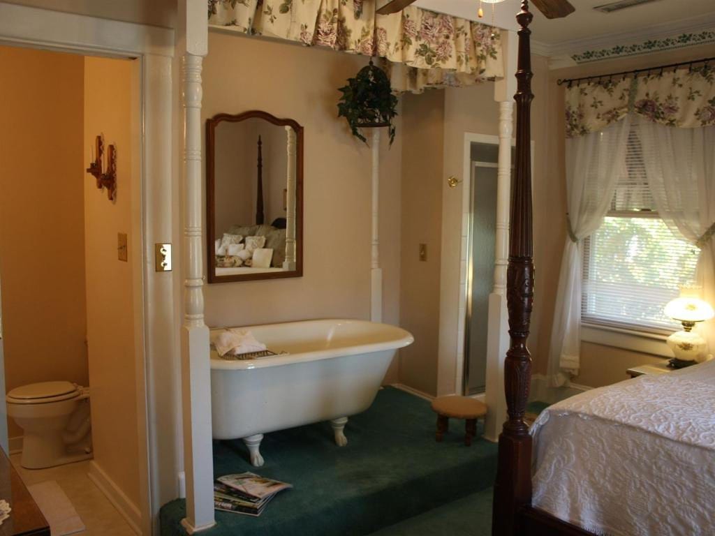 Bathroom with clawfoot tub on a green carpeted platform; bedroom with four-poster bed and window with floral curtains visible.