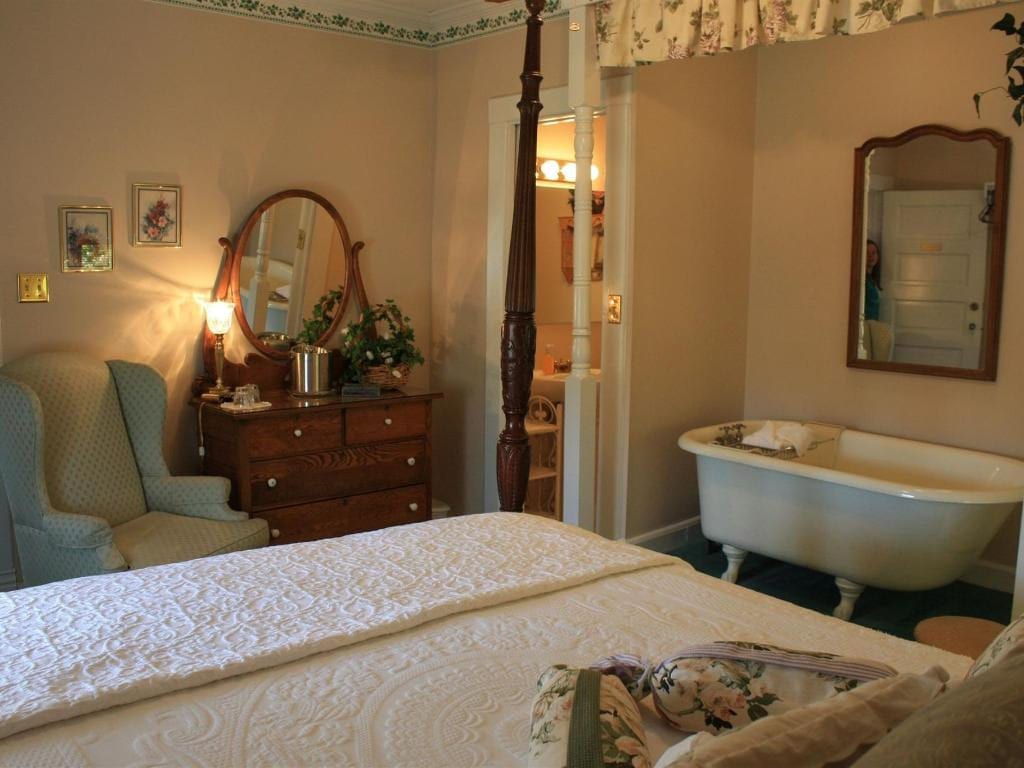 A bedroom with a four-poster bed, armchair, dresser, and a clawfoot bathtub near a doorway to a bathroom.