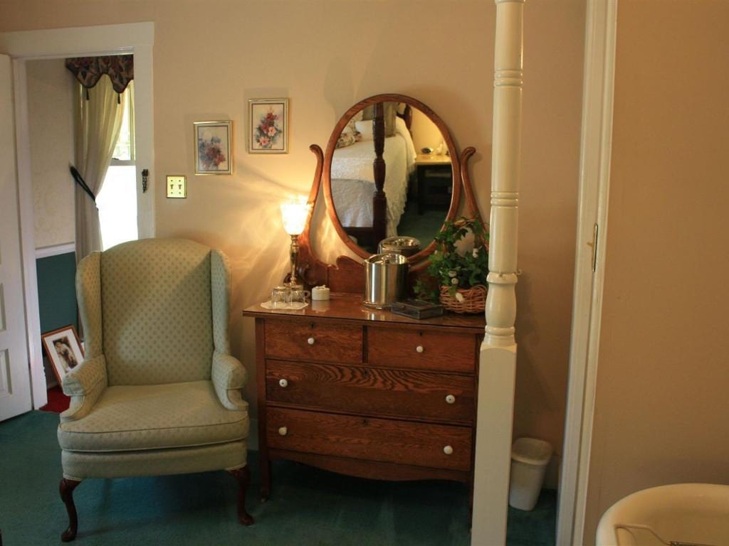 An antique wooden dresser with a mirror, next to a green armchair, in a bedroom. The room has light-colored walls and a window.