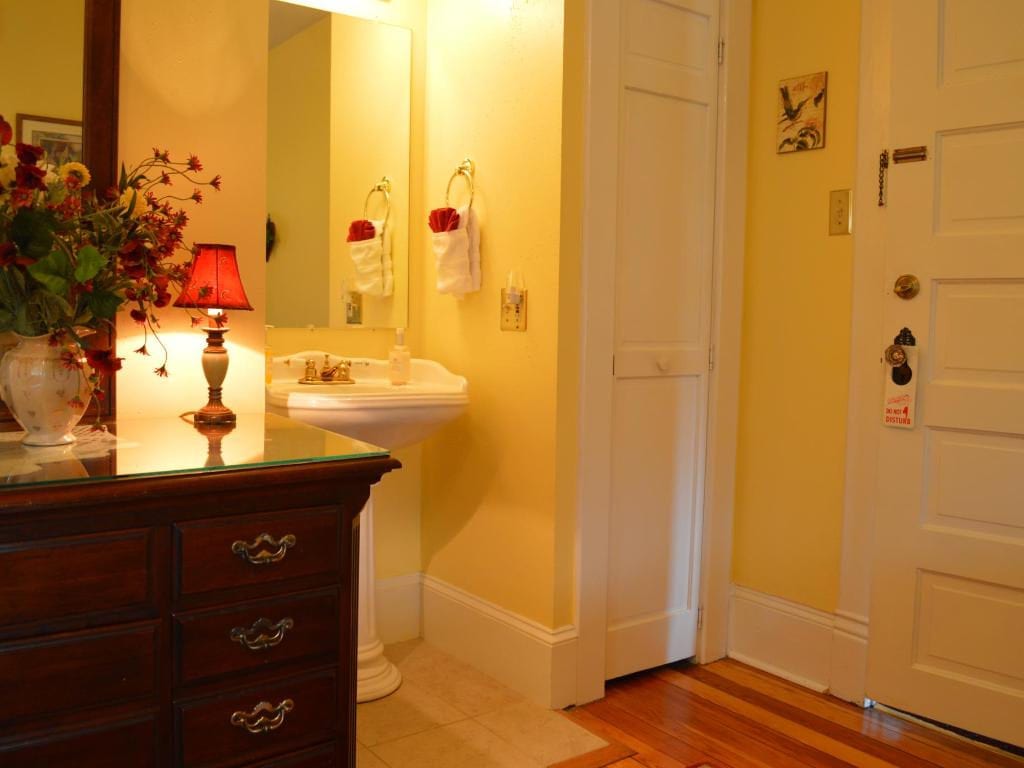 A yellow-walled bathroom with a pedestal sink, a dresser, and a white door. A small lamp sits on the dresser.