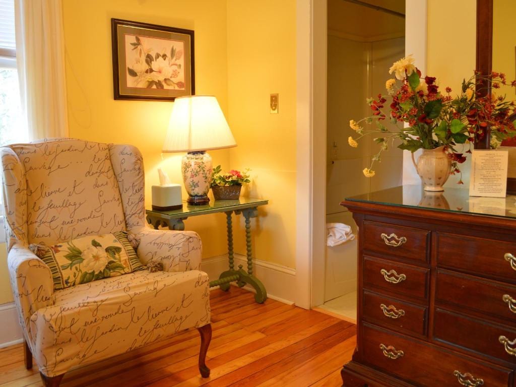 Cozy room with armchair, table, and wooden dresser. Yellow walls, floral decorations, and light-colored patterned armchair.