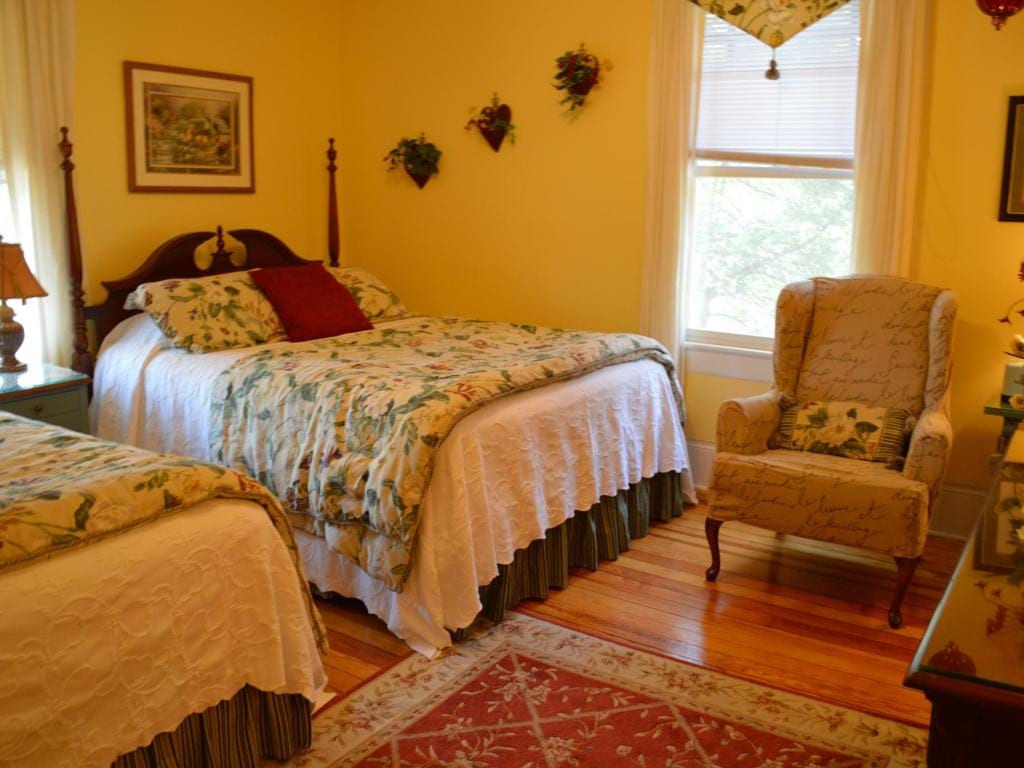 Bedroom with yellow walls, two twin beds, a patterned armchair, and wooden floors.  A window is visible.