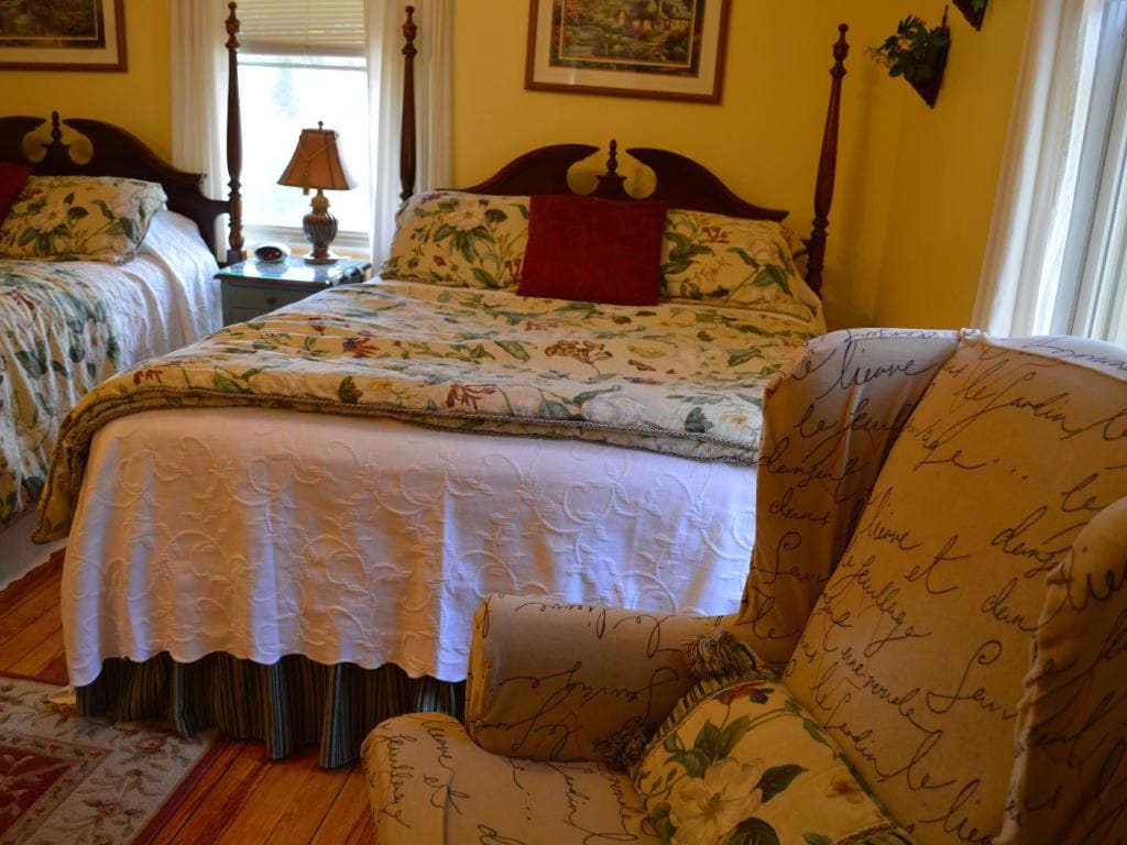 A bedroom with two beds, a floral-patterned quilt, and a patterned armchair. The walls are yellow, and a small rug is on the wooden floor.