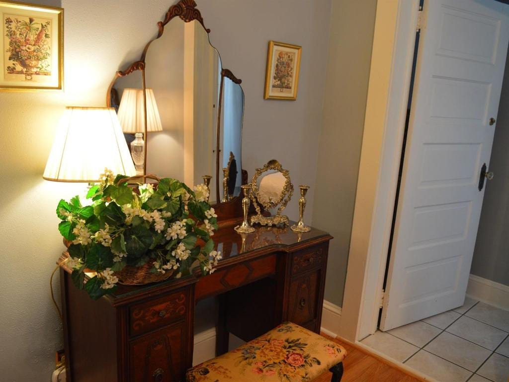 An antique vanity with ornate mirror, lamp, and floral arrangement. Next to a doorway, a patterned stool sits below.