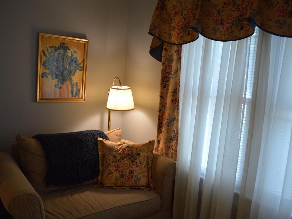 Cozy corner with a chair, lamp, painting, and window with floral curtains. Neutral tones with a pop of color from the artwork and textiles.