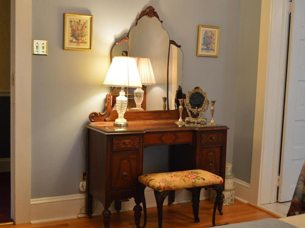 Antique wooden vanity with ornate mirror, two lamps, and a stool against a gray wall.  Two framed pictures hang above the vanity.
