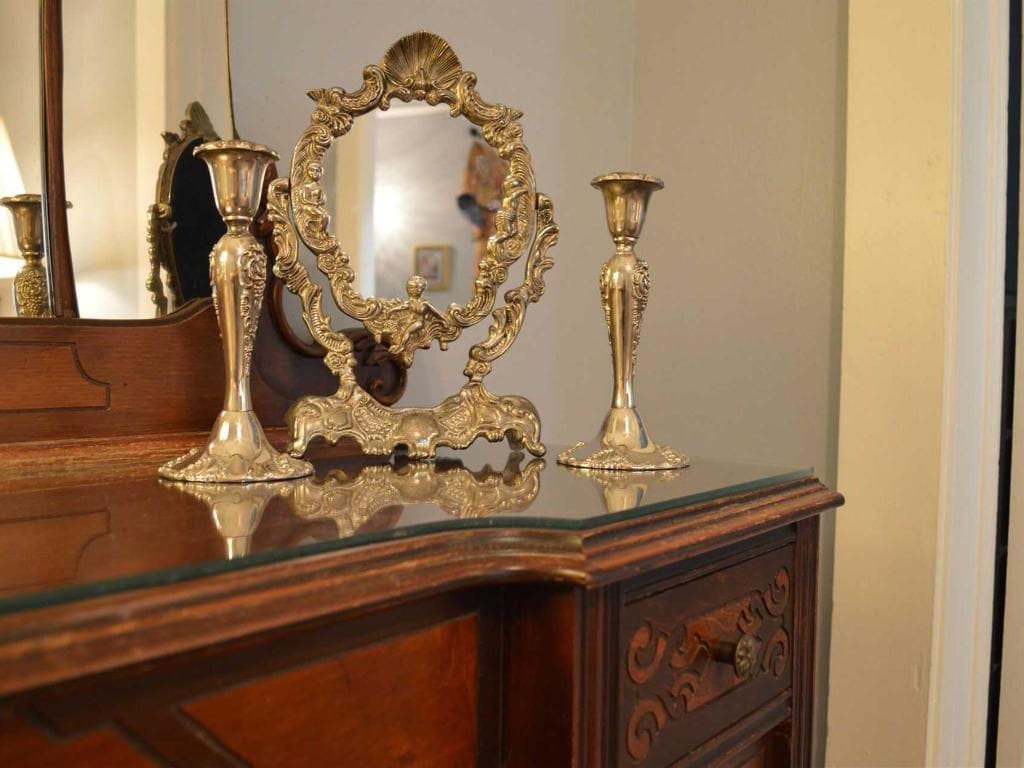 Antique vanity with ornate gold mirror and candlesticks on a wooden dresser. Soft lighting illuminates the scene.