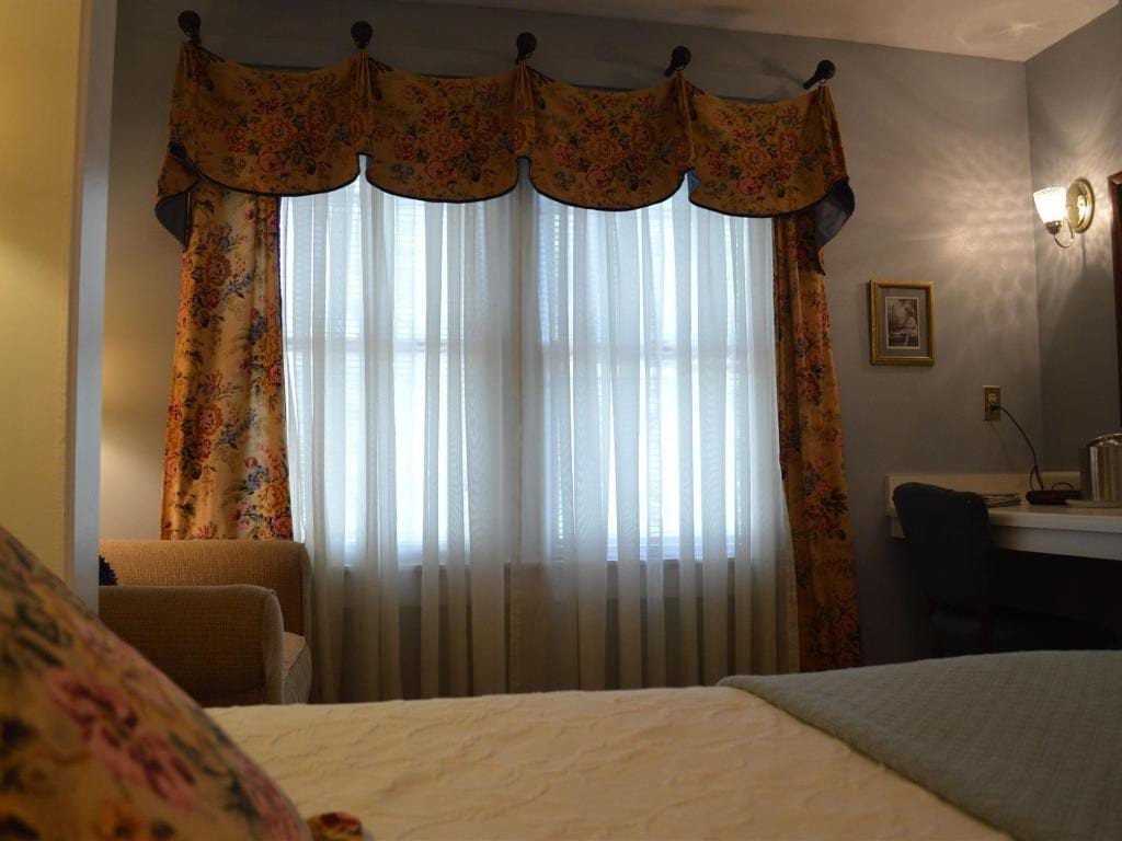 Bedroom with floral curtains and valance, sheer white curtains, and a desk. A bed with a floral pillow is in the foreground.