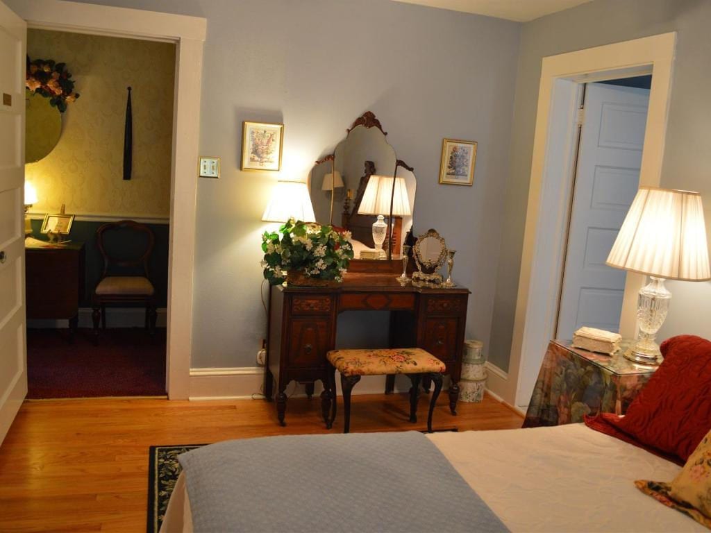 Bedroom with a wooden vanity, floral decor, and a bed. A doorway leads to another room. Light blue walls.