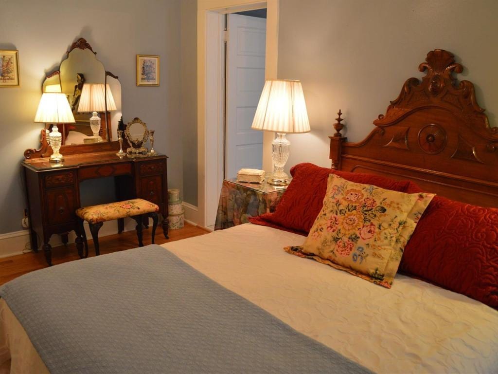Cozy bedroom with a large wooden bed, vanity table, and lamps casting a warm glow. Soft blue walls and red pillows add color.