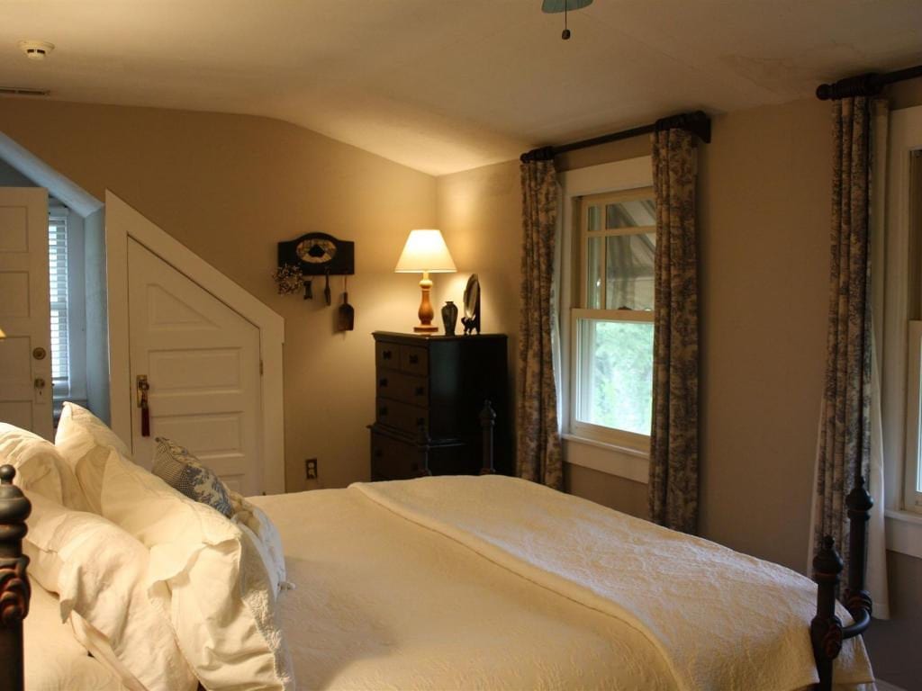 Bedroom with a bed, dresser, lamp, and window with floral curtains. Light-colored walls and a slanted ceiling.
