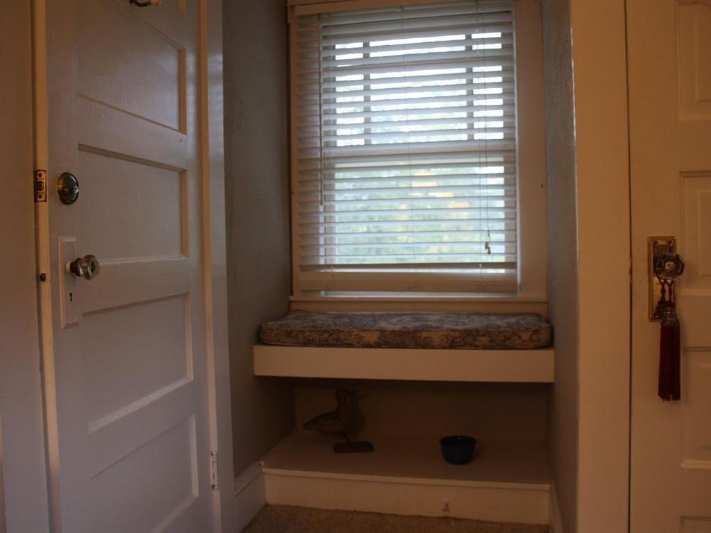 A narrow alcove with a window seat, blind, and a shelf below. A partially open white door and a door with a key hang on either side.