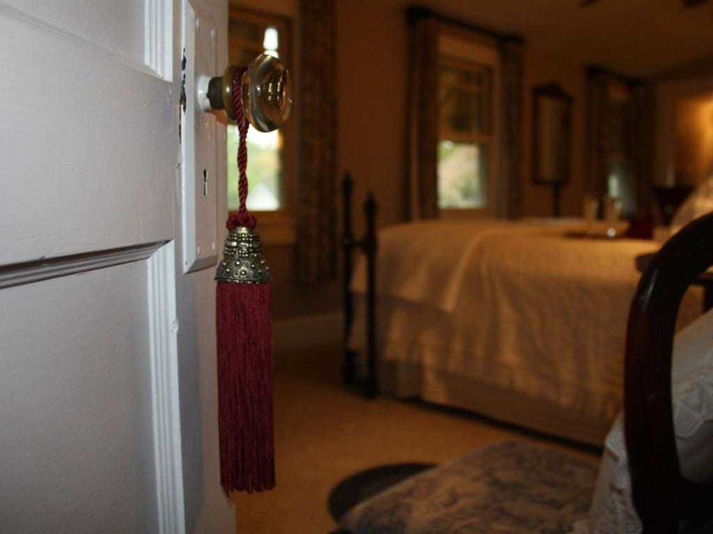 White door ajar, showing a bedroom with a bed and window. A red tassel hangs from the glass doorknob.