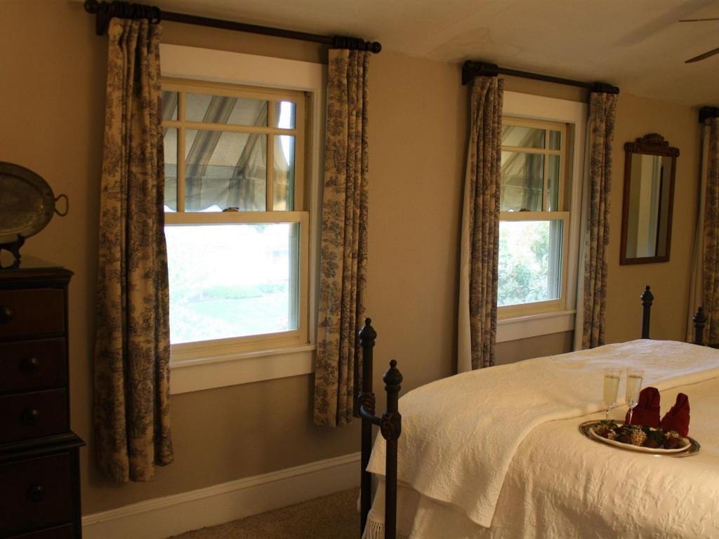 Bedroom with two windows, cream-colored walls, patterned curtains, and a bed with a white cover.