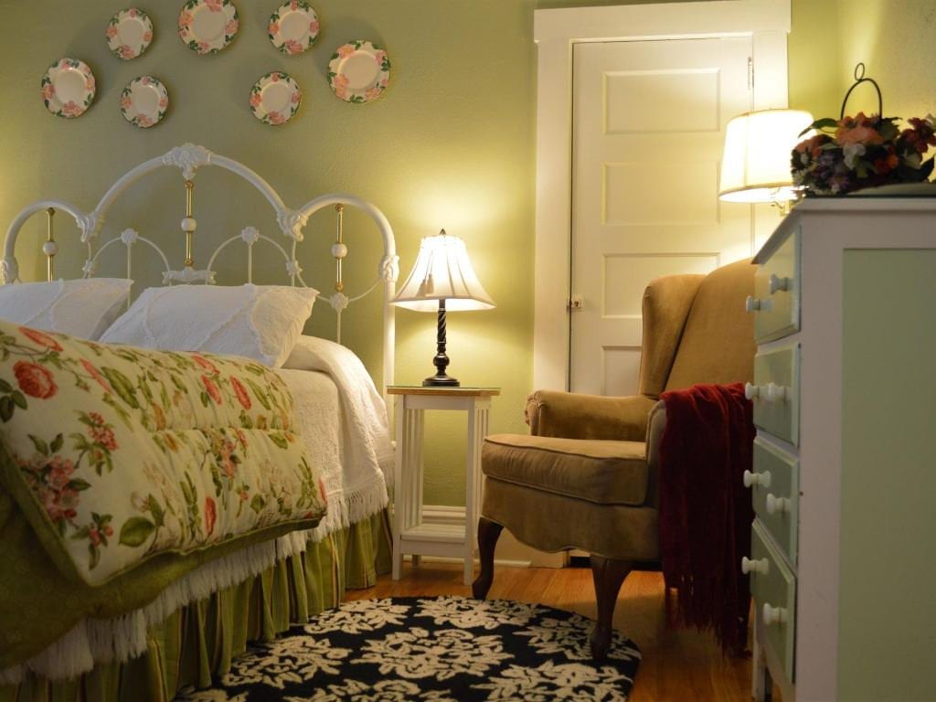 A cozy bedroom with a white bed, floral quilt, and an armchair, lit by lamps, with decorative plates on the wall.