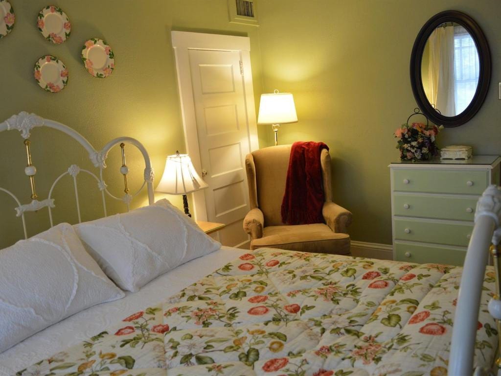 A bedroom with a white wrought-iron bed, a floral quilt, a chair with a red throw, and a mint green dresser.
