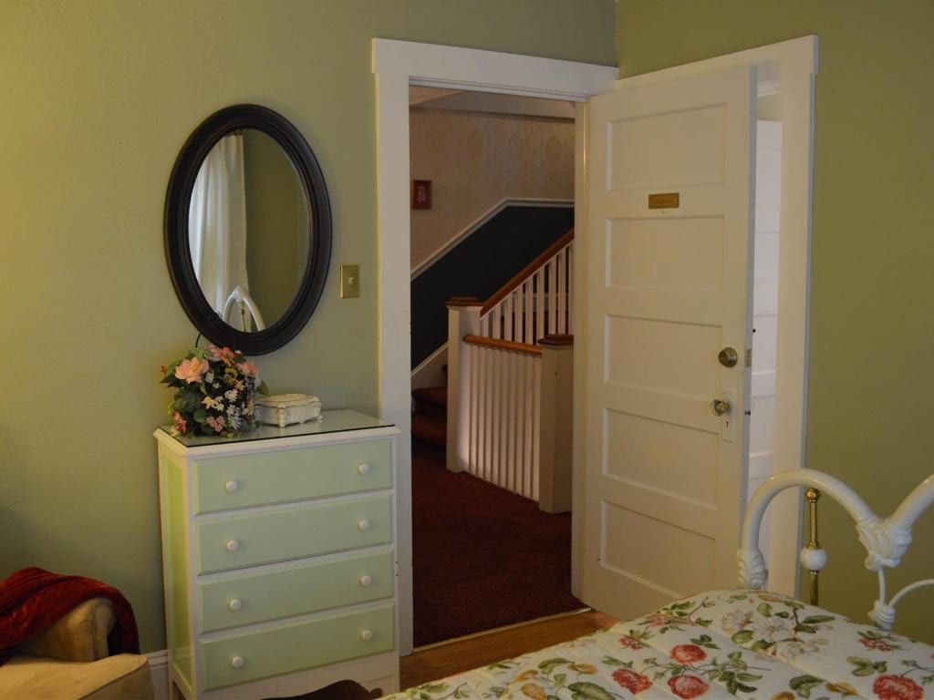 A bedroom doorway reveals a staircase. The room features a chest of drawers with a mirror, and part of a bed.