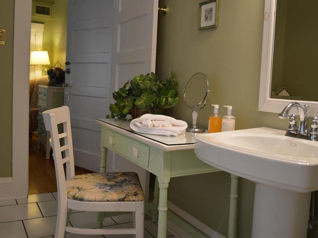 Bathroom with a white pedestal sink, vanity table, and a door leading to a bedroom with a light-colored dresser and lamp.