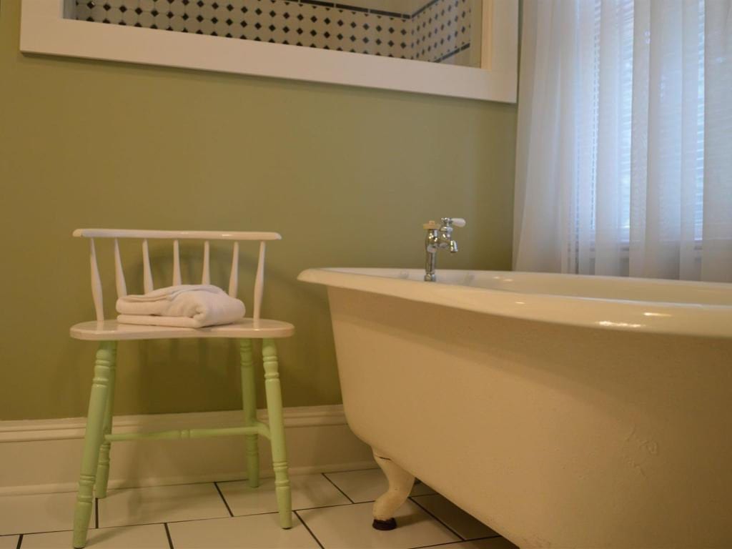 A clawfoot bathtub next to a light green chair with a white towel in a bathroom with light green walls and a white window.