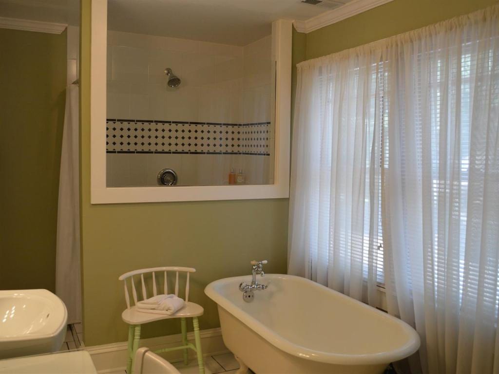 Bathroom with a clawfoot tub, small chair, and a shower enclosed by a glass panel. Light green walls and white sheer curtains.